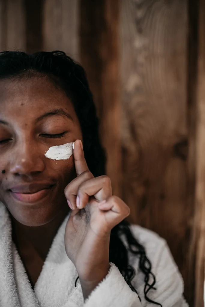 Une femme souriante applique une crème exfoliante blanche sur sa joue en fermant les yeux.
